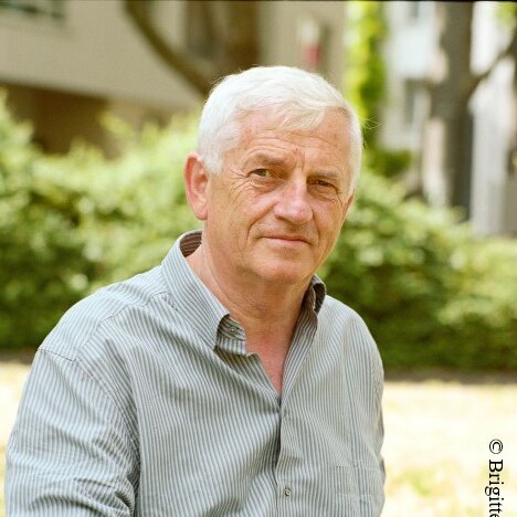 Der Autor Arno Stadler sitzt in einer Wiese und schaut kritisch blickend in die Kamera. 