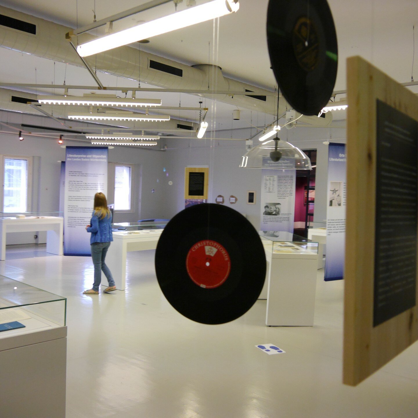 Ein Foto: Ein Ausstellungraum in dem Schallplatten von der Decke hängen. Im Raum sind Vitrinen verteilt. Menschen schauen sich die Ausstellung an. 