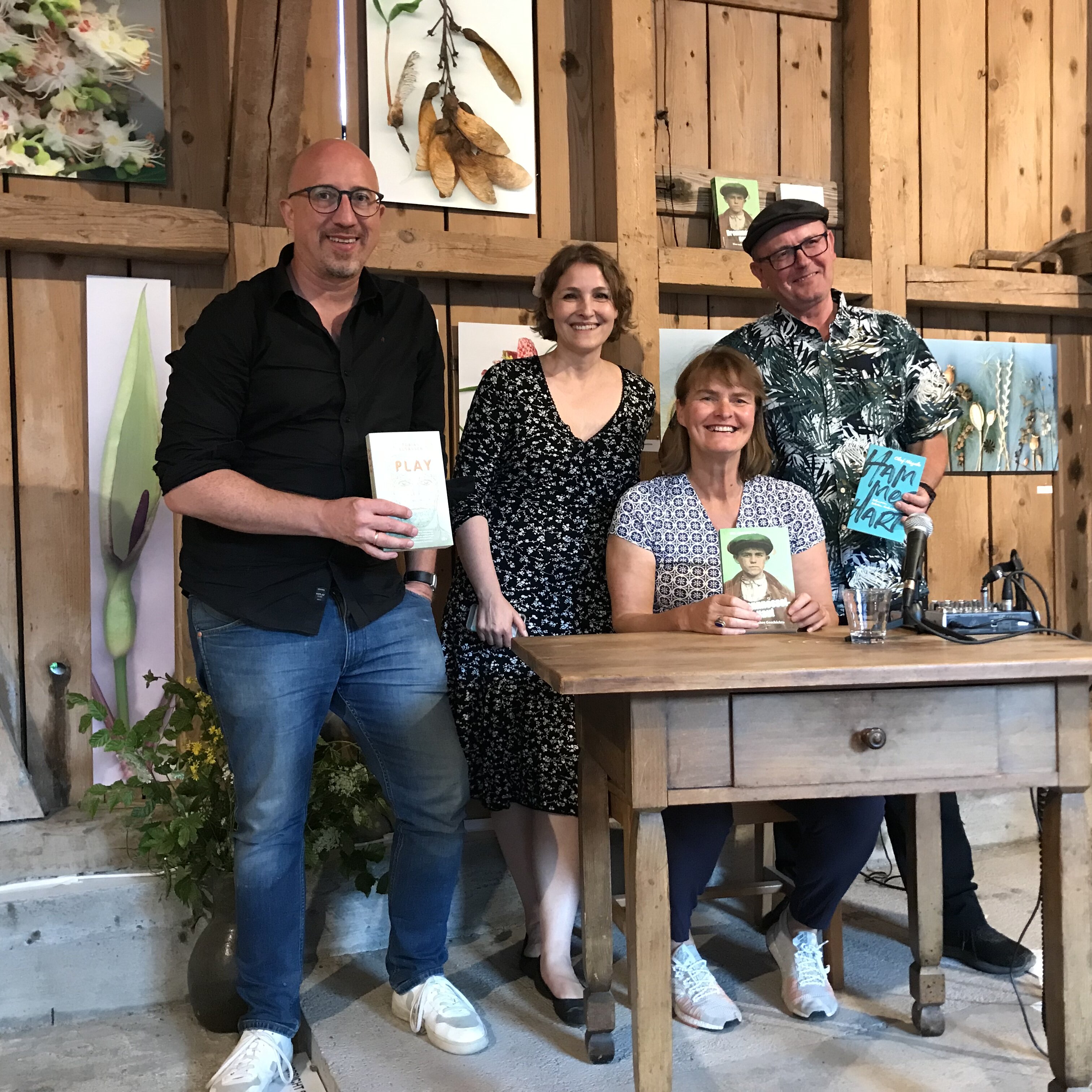 Ein Foto: Ein alter Holztisch mit Schublade an dem eine Frau sitzt. Um sie stehen noch drei Personen vor einer Holzwand. Alle lächeln. 