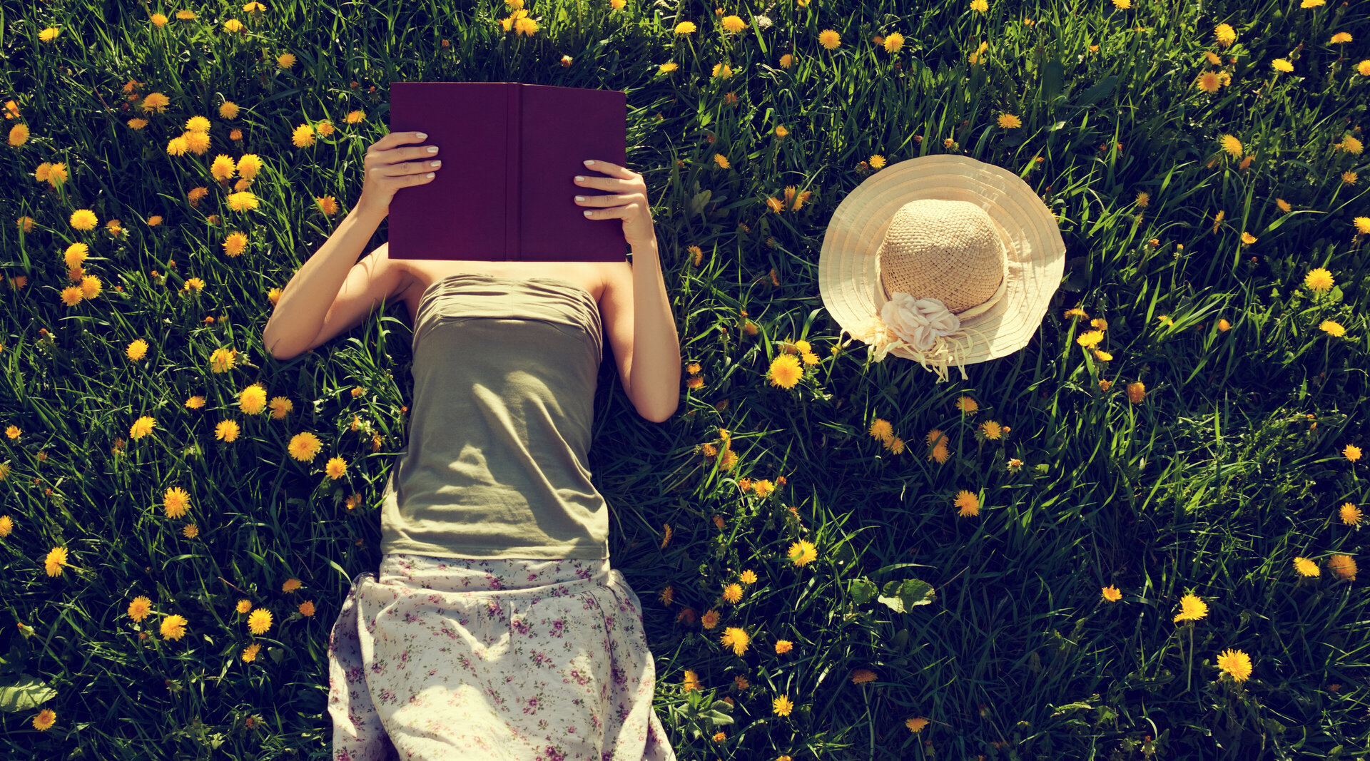 Ein Stimmungsbild. Eine Frau liegt in einer blühenden Wiese und liest. Dabei hat sie das Buch vor ihr Gesicht gehalten. Neben ihr liegt ein Strohhut im Gras.